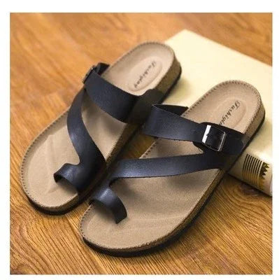 Black leather cross-strap sandals with toe loop on wooden floor next to rolled paper
