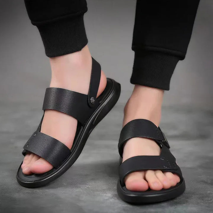 Black leather open-toe mens sandals with adjustable straps worn with black pants on gray floor