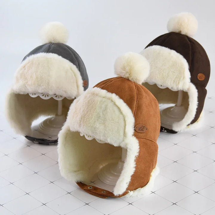 Three warm winter trapper hats in brown, gray, and black with white faux fur lining and pom poms