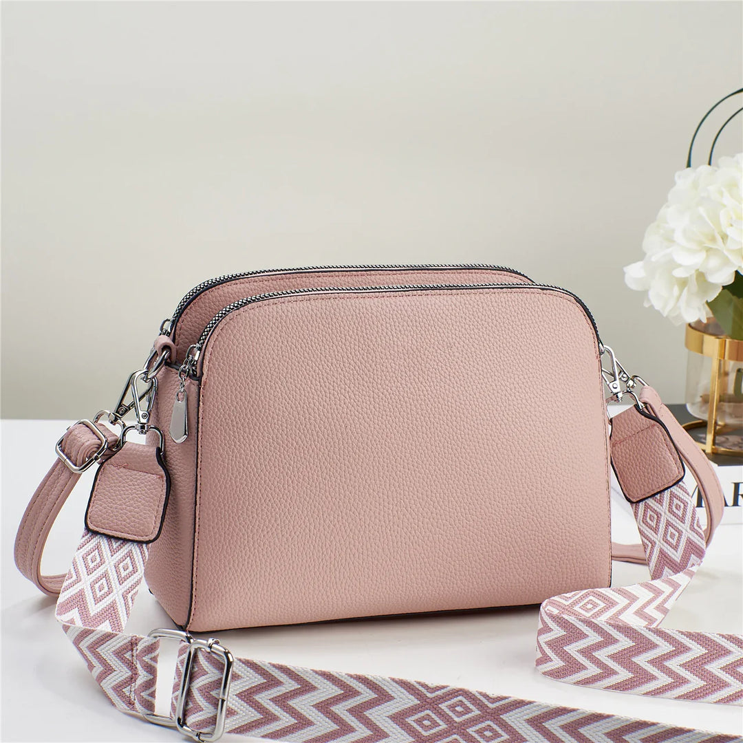 Blush pink textured leather crossbody bag with dual zippers and adjustable patterned shoulder strap on white surface