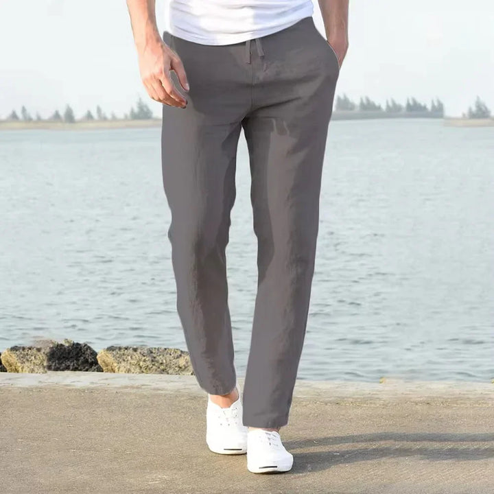 Man wearing gray jogger pants and white sneakers walking by a waterfront with rocks and distant trees