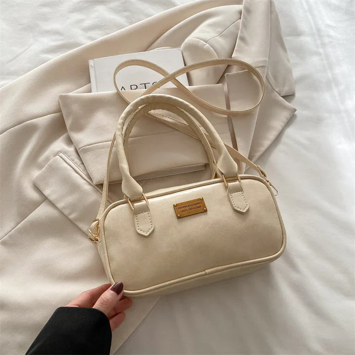 Beige leather handbag with gold label and double handles on cream fabric with folded clothes and art book