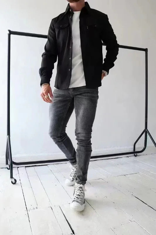 Man wearing black jacket, white t-shirt, grey jeans, and white sneakers posing indoors