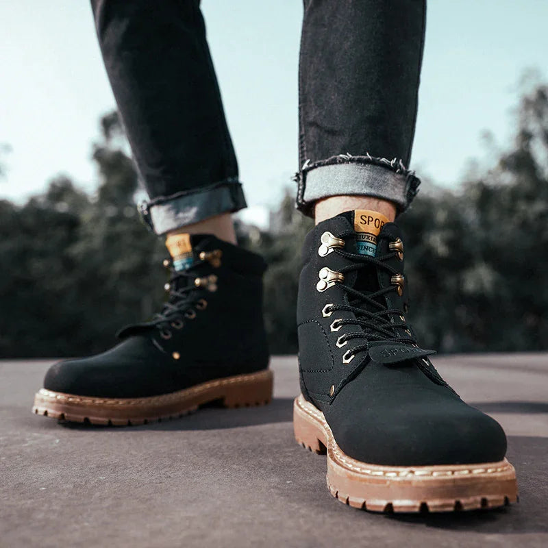 Black leather men's hiking boots with brown soles and gold eyelets worn with rolled-up jeans outdoors