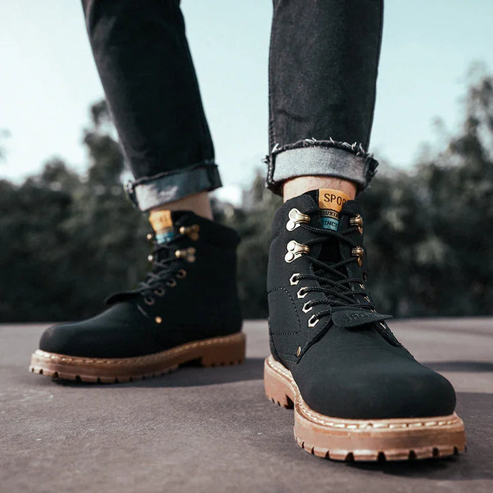 Black leather men's hiking boots with brown soles and gold eyelets worn with rolled-up jeans outdoors