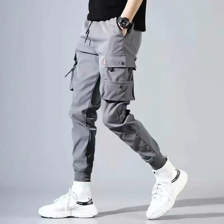 Man wearing grey cargo jogger pants with multiple pockets and white sneakers against grey background