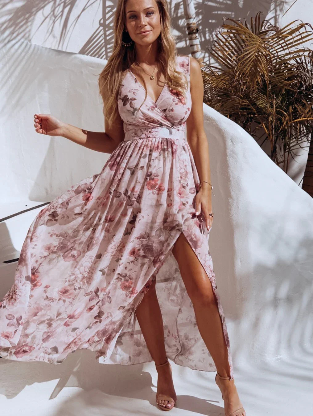 Woman in sleeveless pink floral maxi dress with thigh slit standing against white wall with shadows