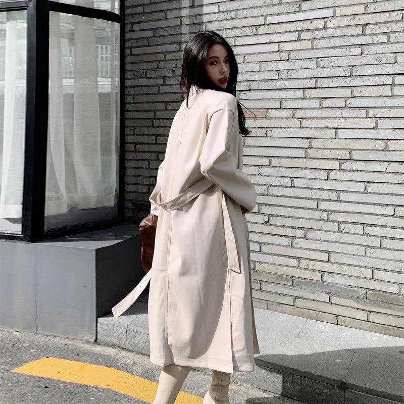Woman wearing beige long trench coat walking beside brick wall with window and curtain