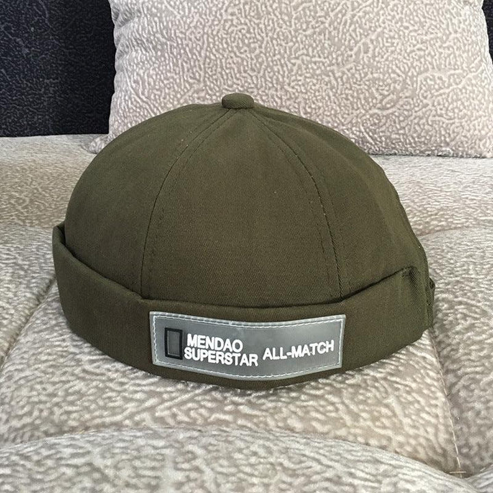 Olive green beanie hat with folded edge and label reading MENDÃO SUPERSTAR ALL-MATCH on textured beige fabric background