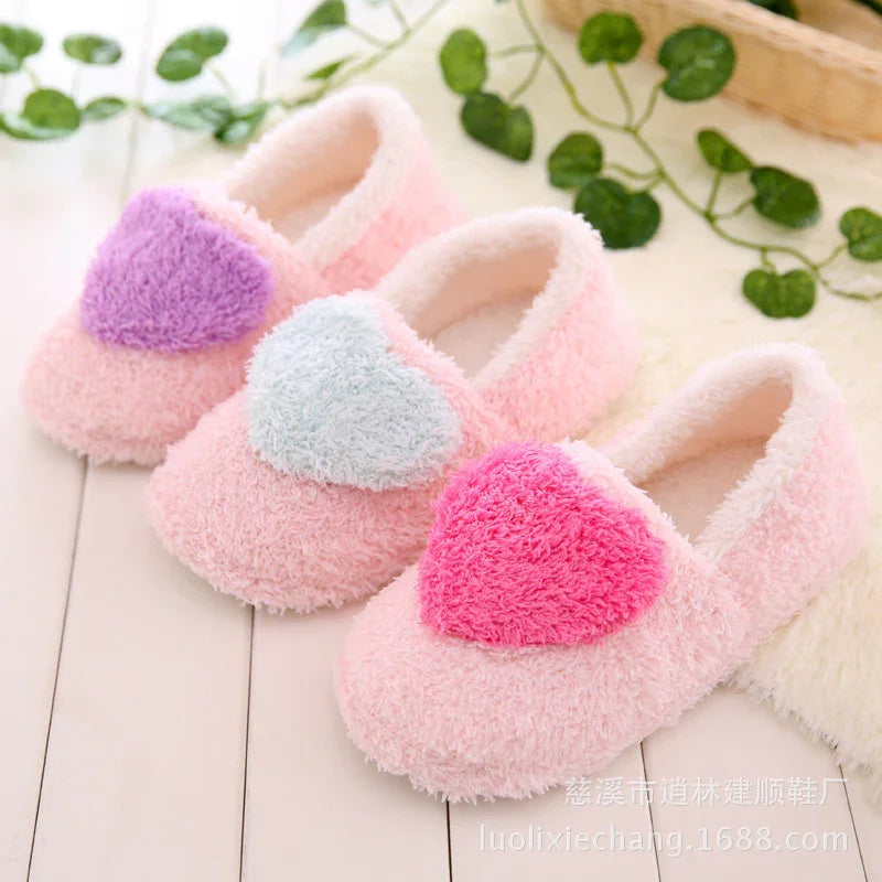 Three pairs of pink fuzzy slippers with large colorful heart patches in purple, light blue, and pink on white floor with green leaves