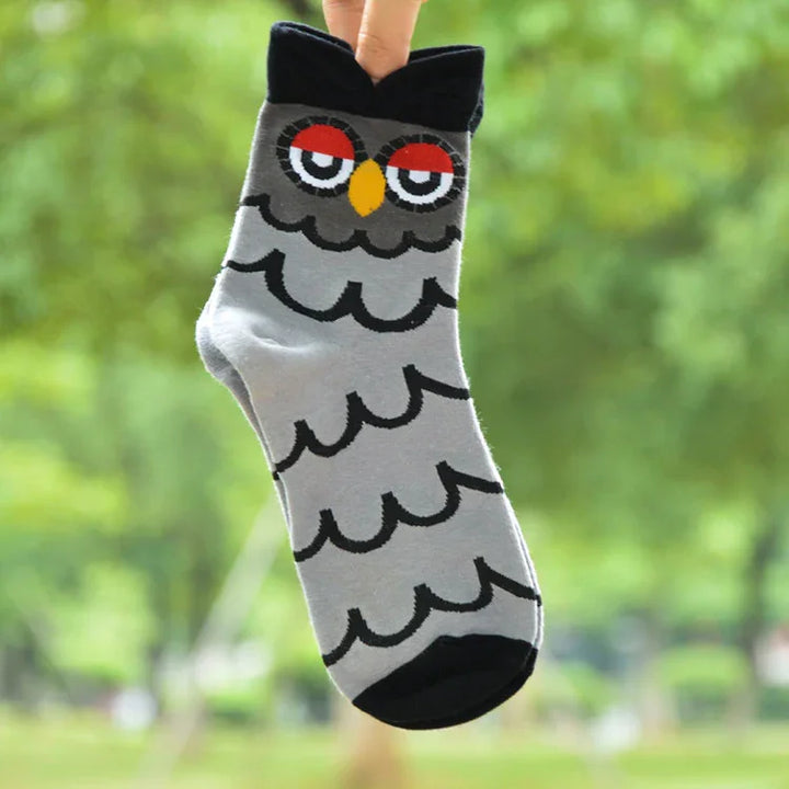 Gray sock featuring a cartoon owl face with red eyelids and yellow beak, held up against a blurred green outdoor background