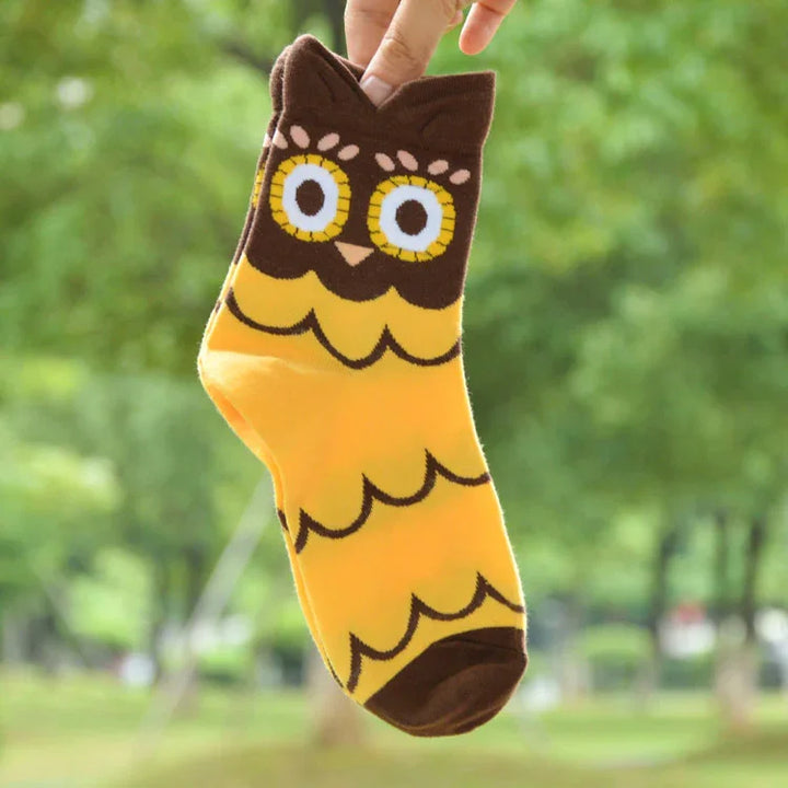 Hand holding a yellow and brown sock with a cute owl face and scalloped pattern against blurred green outdoor background