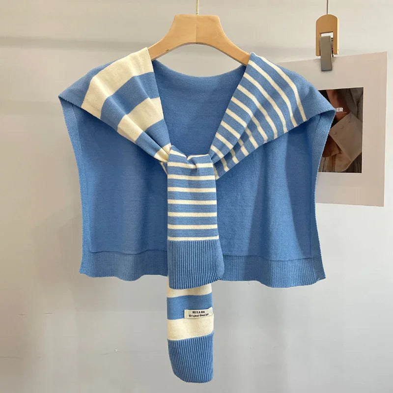 Blue knitted sleeveless cape with striped scarf draped over wooden hanger on beige wall