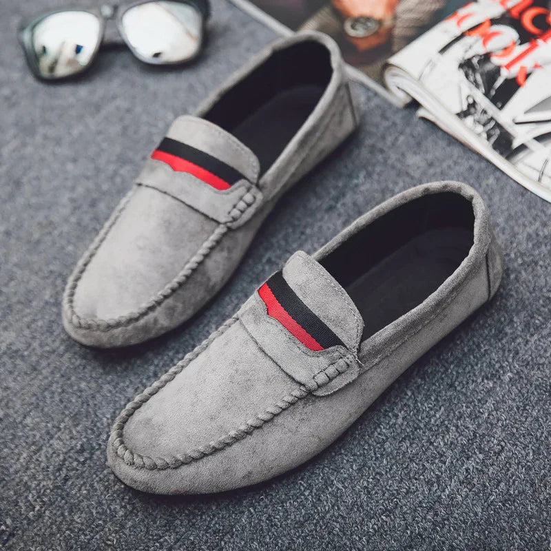 Gray suede loafers with black and red stripe detail on top, styled with sunglasses and a folded magazine
