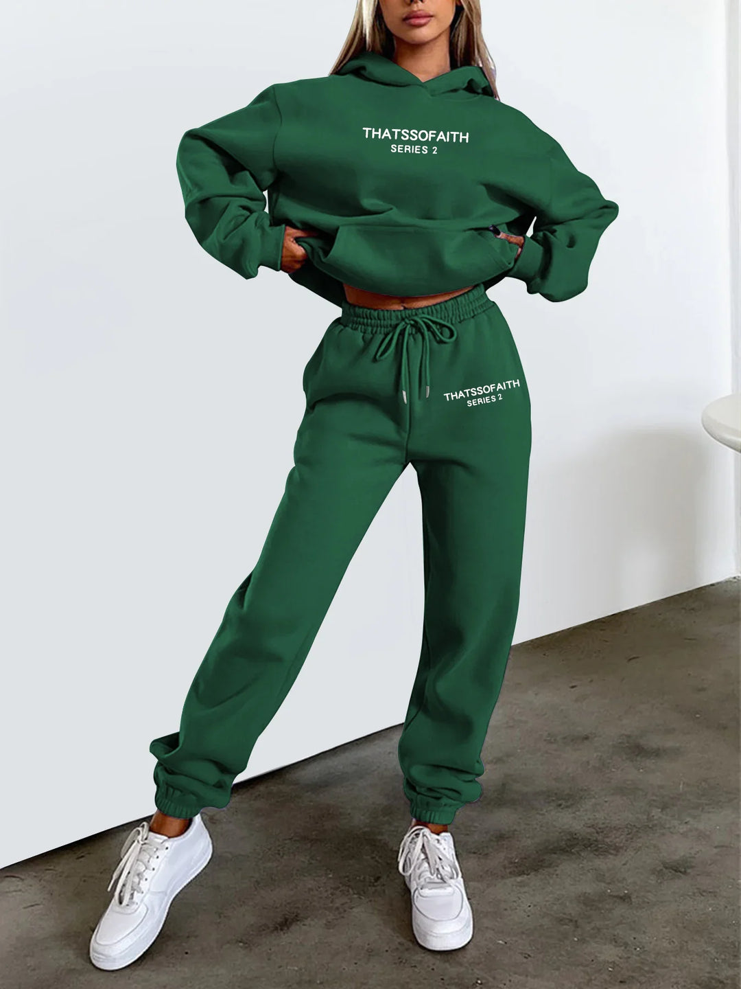 Woman wearing green hoodie and sweatpants set with THATSSOFAITH Series 2 text, white sneakers, casual outfit