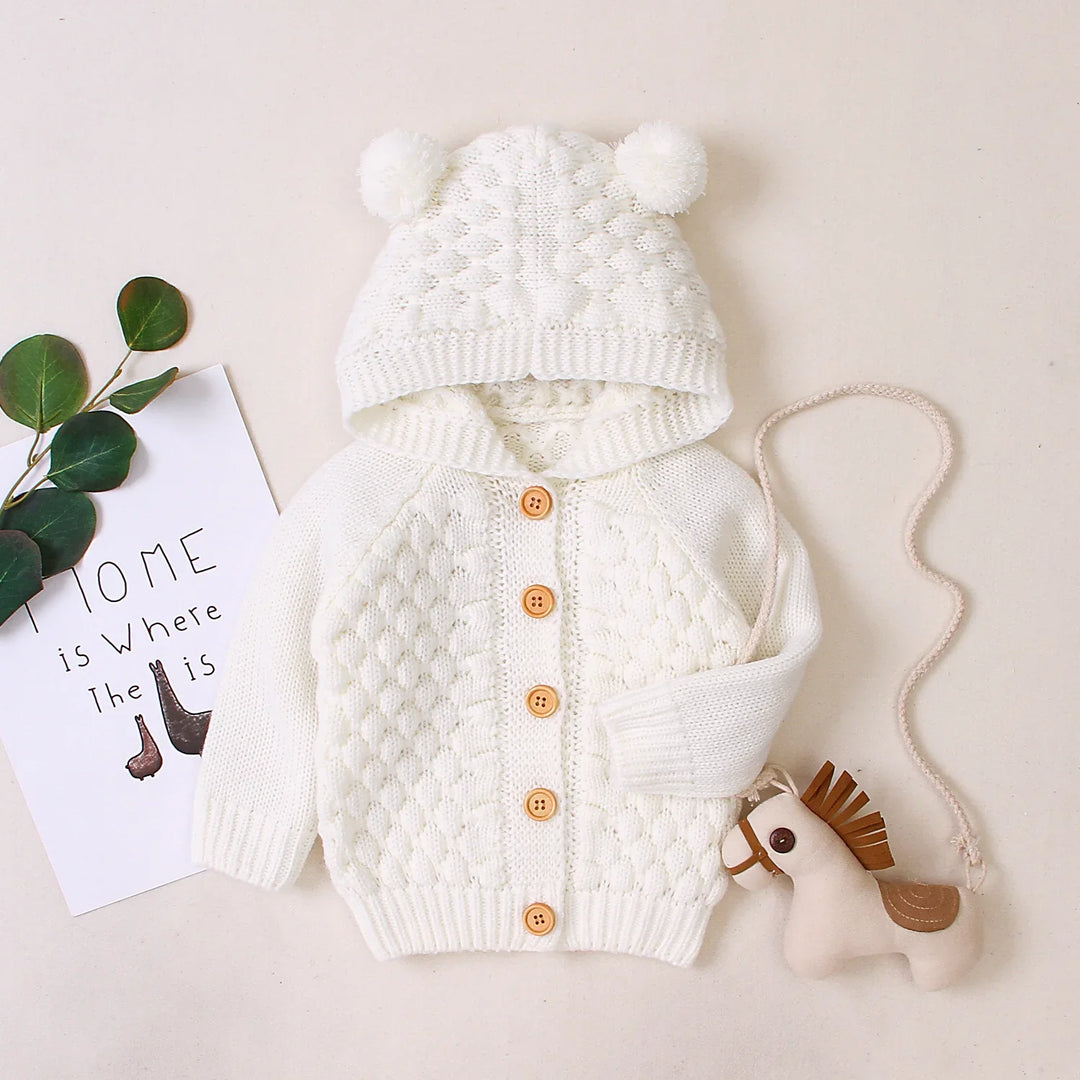 White knitted baby cardigan with hood and pom-poms, wooden buttons, toy horse, and decorative leaves