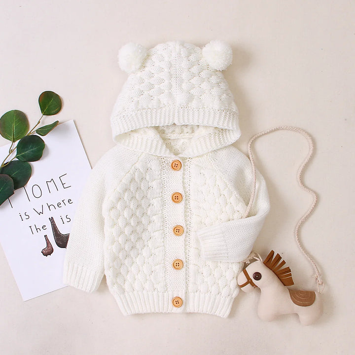 White knitted baby cardigan with hood and pom-poms, wooden buttons, toy horse, and decorative leaves