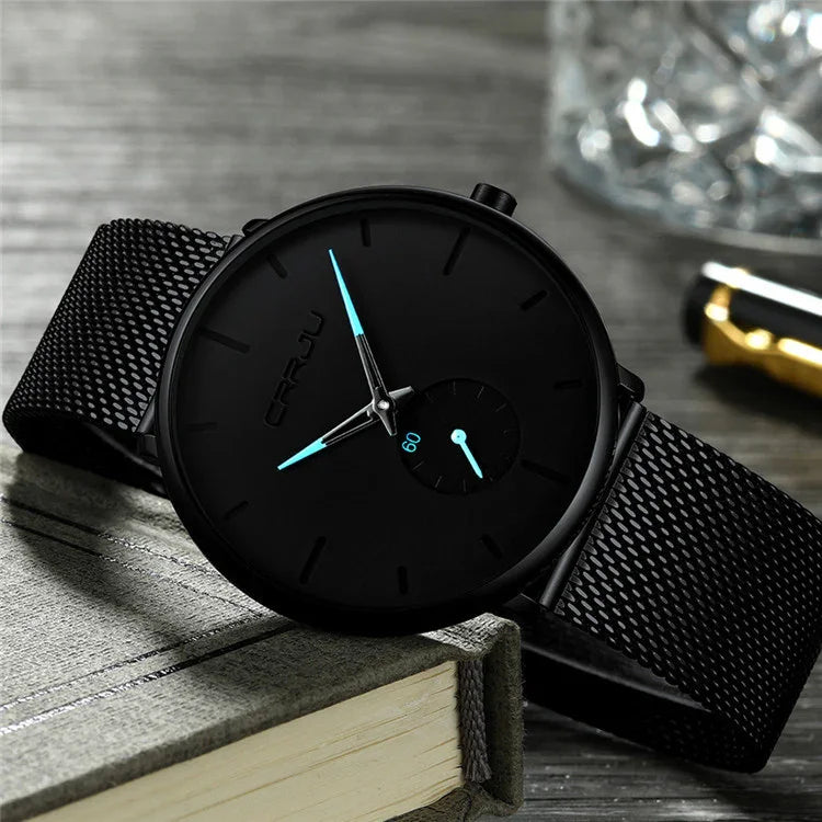 Black minimalist wristwatch with blue hands and mesh strap resting on a closed book on wooden surface