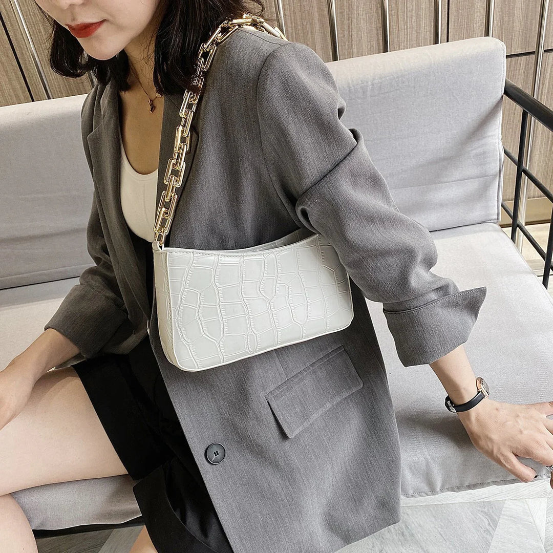 Woman wearing gray blazer and black skirt sitting on gray sofa with white crocodile pattern shoulder bag and gold chain strap