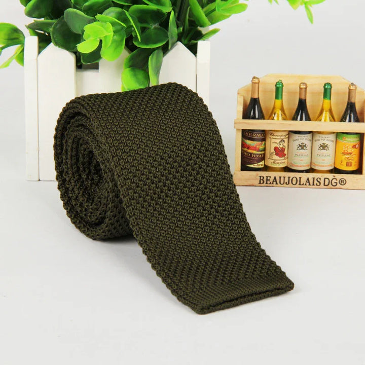 Rolled olive green knitted tie on white surface with small plant and decorative wine bottles in background