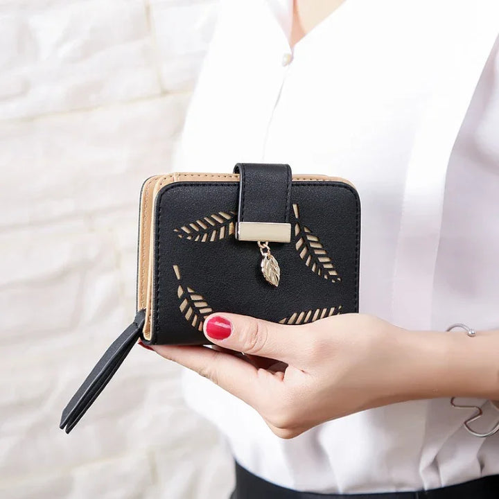 Black leather wallet with leaf cutout design and gold leaf charm held by woman with red nail polish