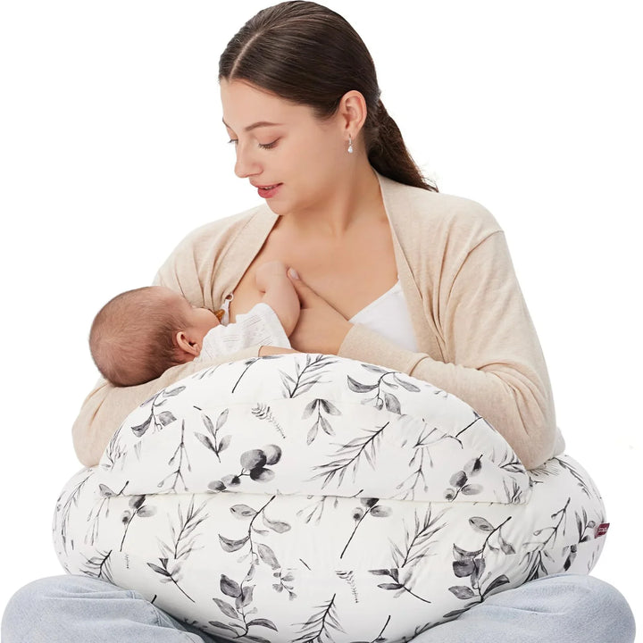 Mother breastfeeding baby using white floral nursing pillow on lap, beige cardigan, white background
