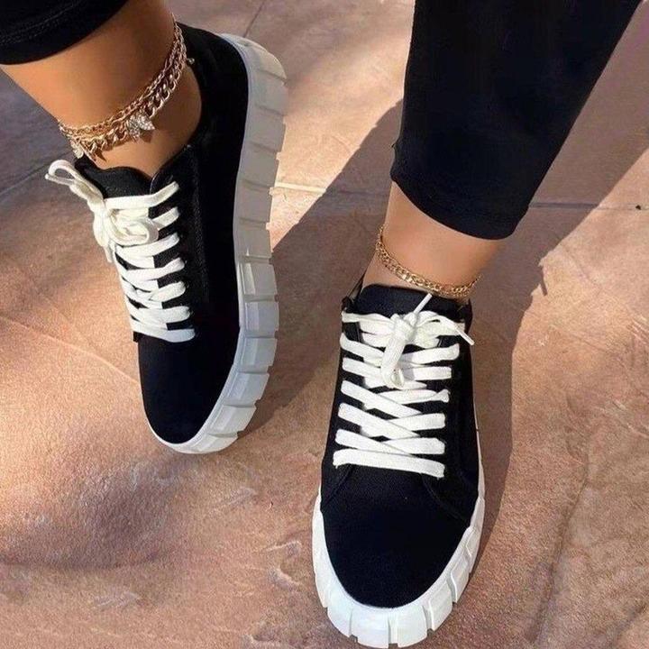 Black sneakers with thick white soles and white laces worn with gold anklets on a textured floor