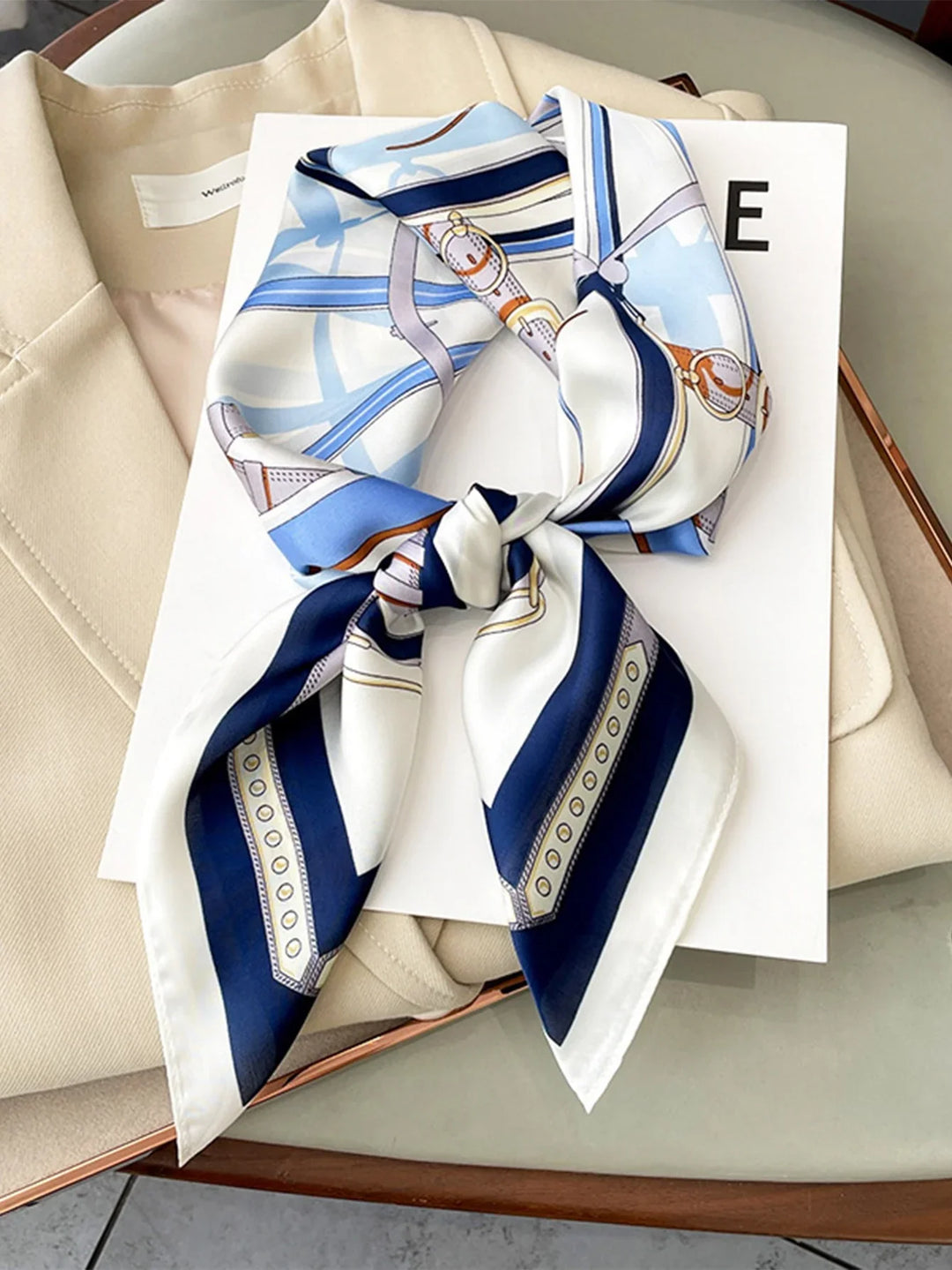 Silk scarf with blue, white, and orange pattern on beige blazer and white book on glass table