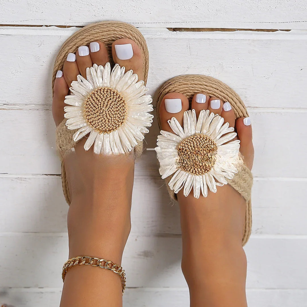 Feet wearing beige sandals with large white daisy decorations and white pedicure on wooden floor