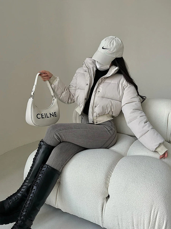 Woman wearing beige puffer jacket, gray leggings, black knee-high boots, white Nike cap, holding white CEILNE handbag, sitting on white textured sofa