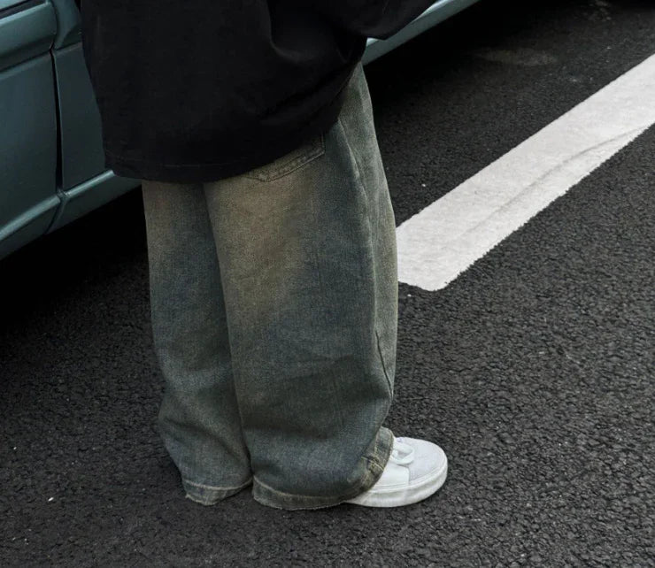 Person wearing loose gray denim jeans and white sneakers standing on asphalt near a teal car