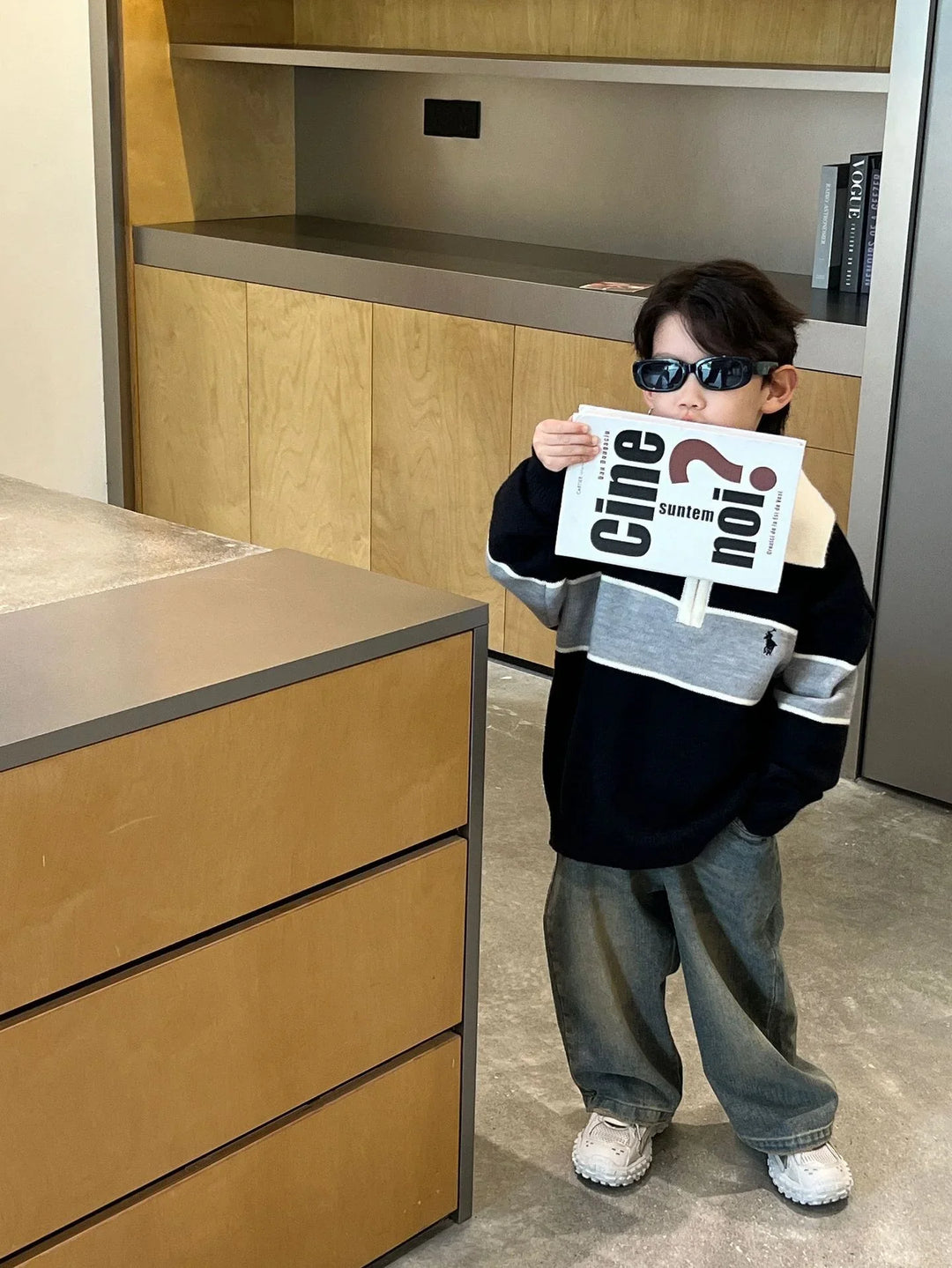 Young boy wearing sunglasses, black and gray sweater, gray pants, holding a large book in a modern kitchen