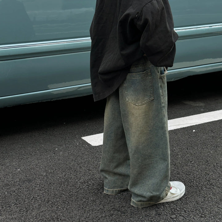 Person wearing black shirt, wide-leg faded jeans, and white sneakers standing on asphalt near light blue vehicle
