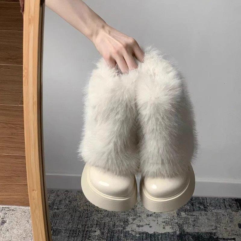 Hand holding a pair of white furry platform boots with glossy toe caps on a gray carpeted floor