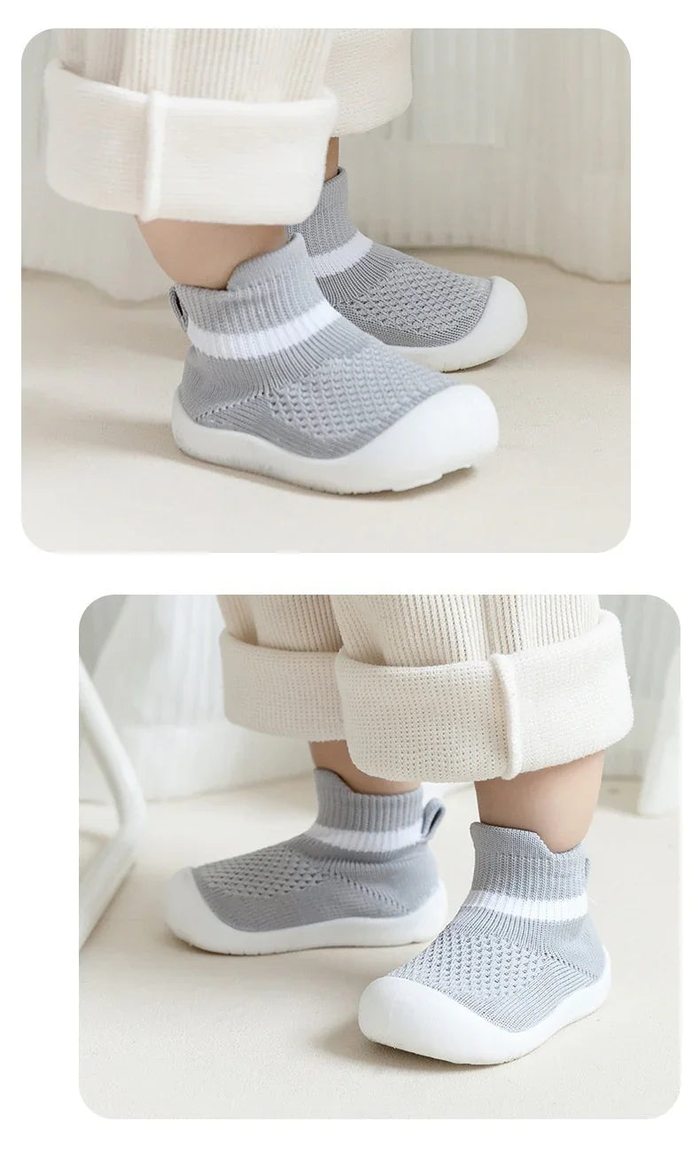 Baby wearing gray knit sock shoes with white soles and cuffed cream pants indoors