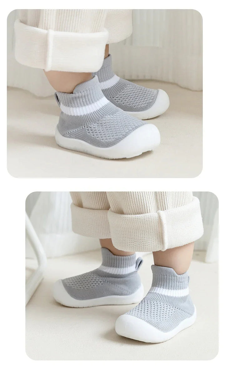 Baby wearing gray knit sock shoes with white soles and cuffed cream pants indoors