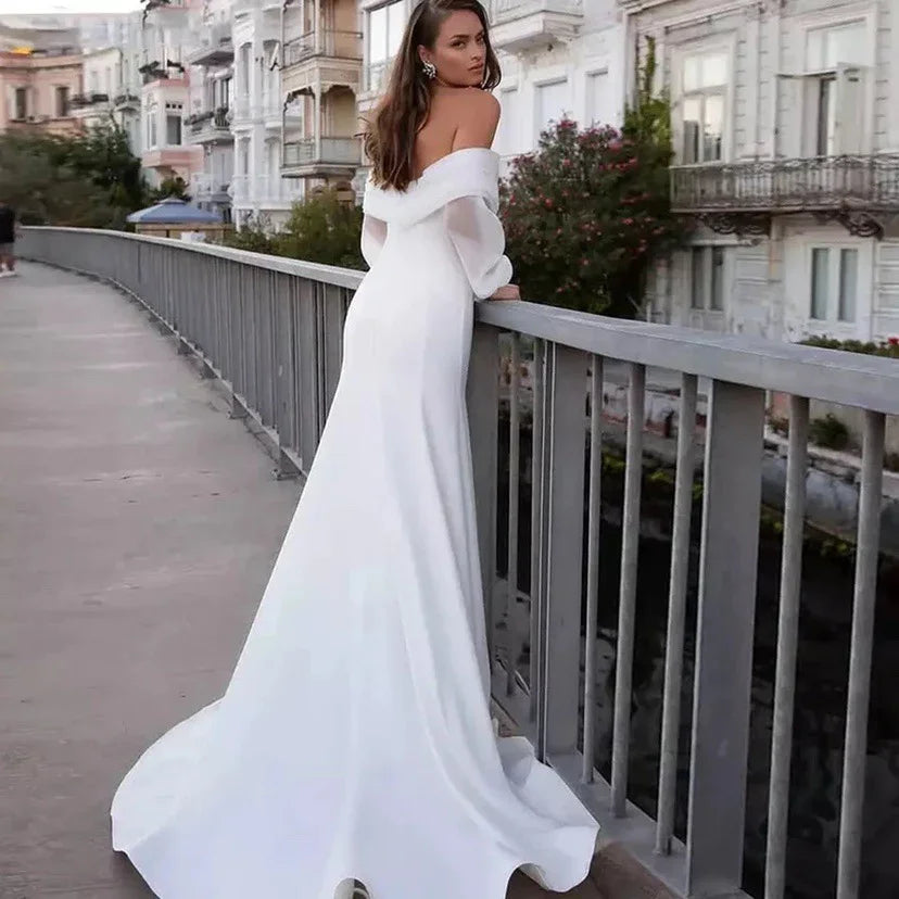 Woman in elegant off-shoulder white wedding dress standing on urban balcony with historic buildings