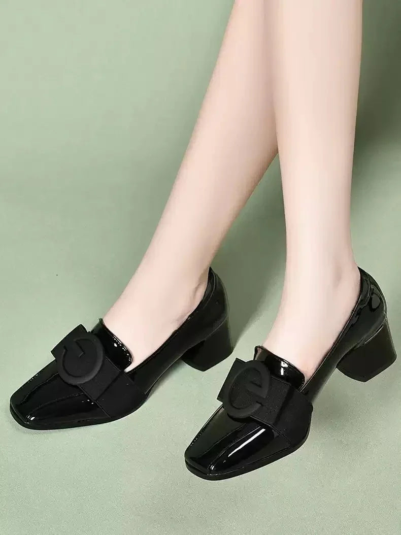 Glossy black block heel shoes with large bow accent worn on feet against green background