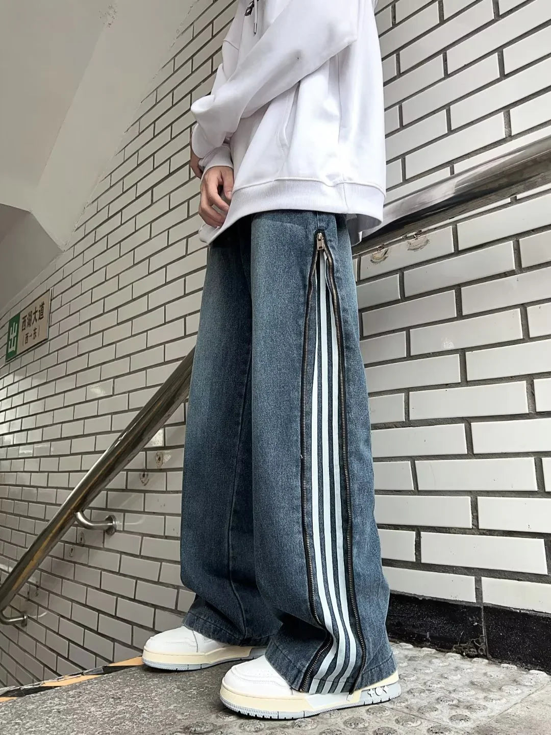 Person wearing white hoodie and wide-leg jeans with side zippers and stripes, standing near tiled wall