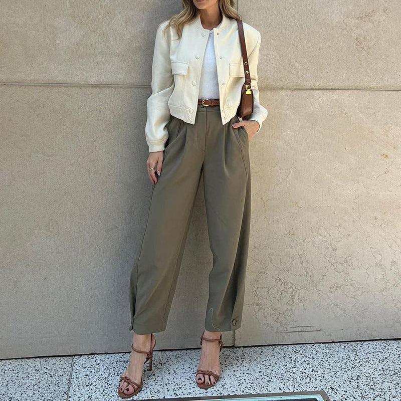 Woman wearing white cropped jacket, olive high-waisted trousers, brown belt, and strappy heels by beige wall