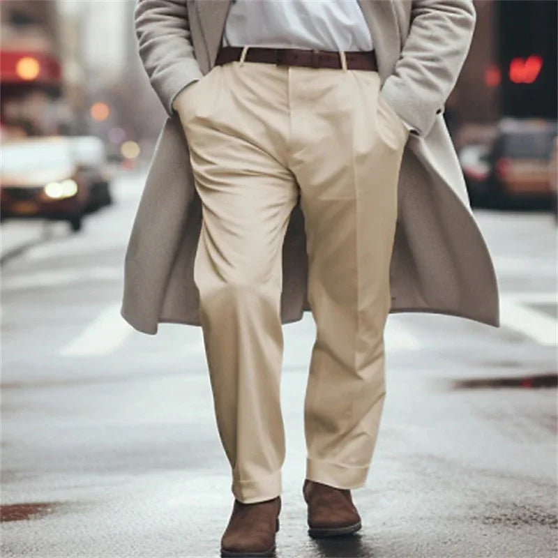 Man walking on city street wearing beige pants, brown belt, brown shoes, and a long light coat