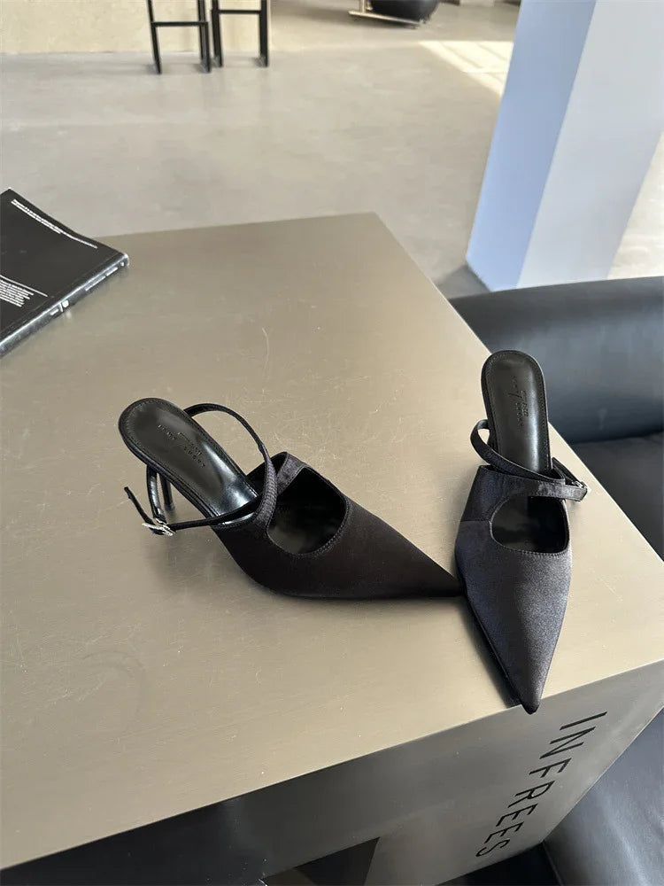 Pair of black pointed-toe high-heeled mules with straps on tan square table in modern indoor setting