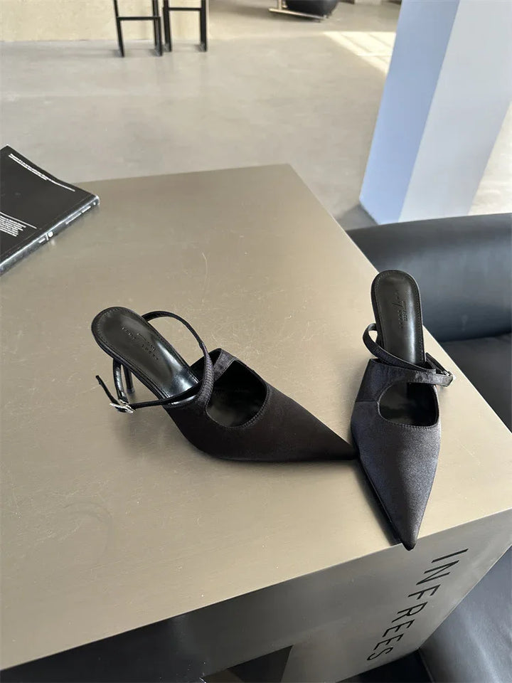 Pair of black pointed-toe high-heeled mules with straps on tan square table in modern indoor setting