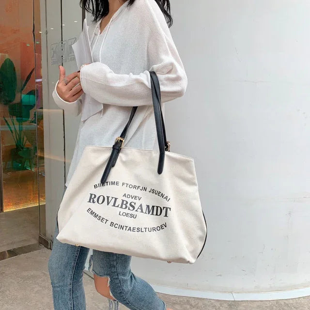 Woman in white sweater carrying beige canvas tote bag with black straps and black text print