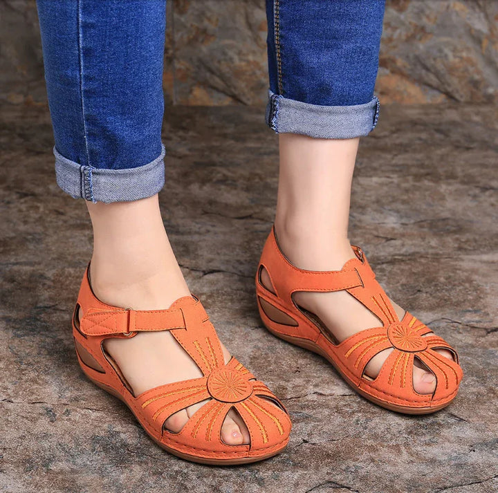 Women's orange casual sandals with floral cutout design worn with rolled-up blue jeans on stone floor