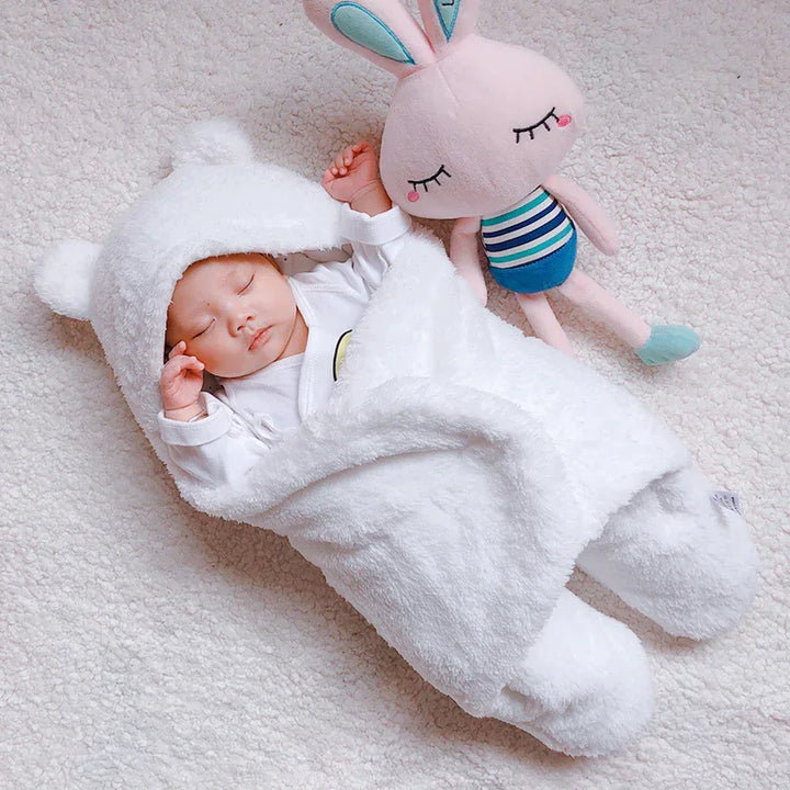 Sleeping baby wrapped in soft white bear hooded blanket lying on plush white surface with pink plush bunny toy