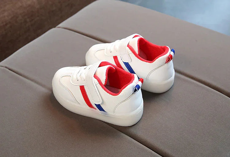 White toddler sneakers with red and blue stripes and red inner lining on brown surface