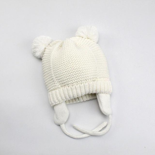 White knitted baby hat with ear flaps and pom-poms on top on a light background