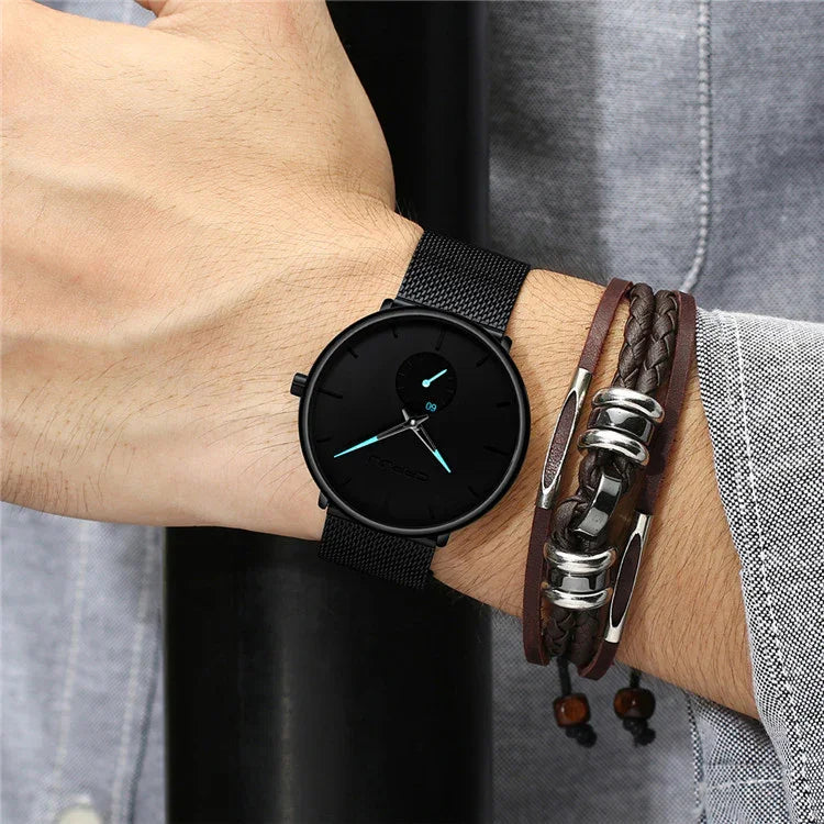 Black minimalist wristwatch with blue hands and black mesh band on wrist, paired with braided leather bracelet