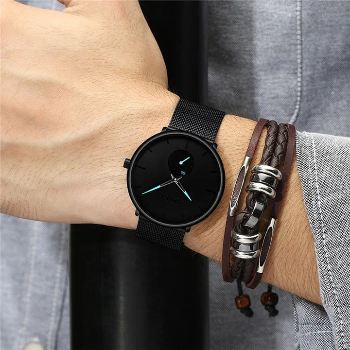 Black minimalist wristwatch with blue hands and black mesh band on wrist, paired with braided leather bracelet